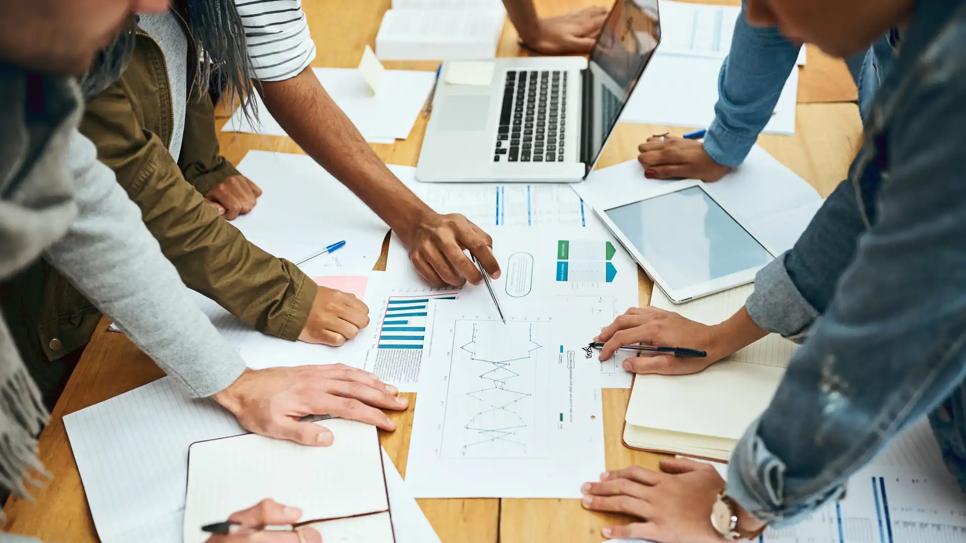 People leaning around a table with a computer, tablet, and papers looking at charts