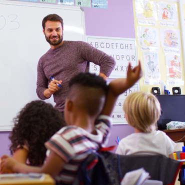 Male teacher in front of classroom with young student raising hand
