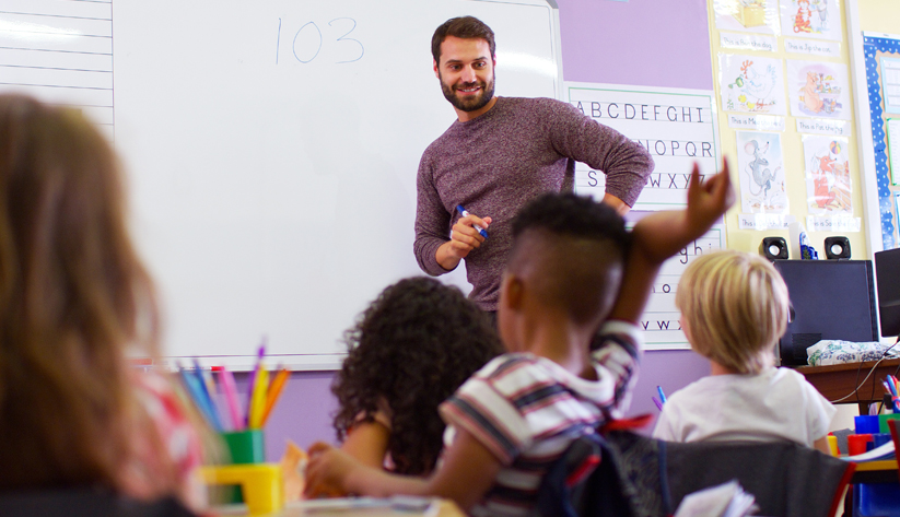Male teacher in front of classroom with young student raising hand