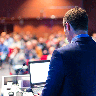 viewer standing behind person seated at podium with audience out of focus in background
