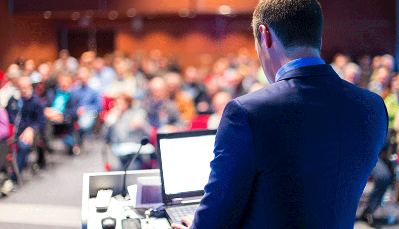 viewer standing behind person seated at podium with audience out of focus in background