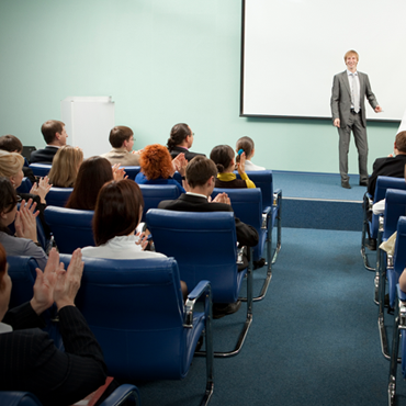 man standing on stage while audience claps as viewed from behind audience