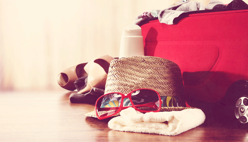 photograph of a summer hat, sunglasses, shoes, and other travel gear on the floor next to a redsuitcase