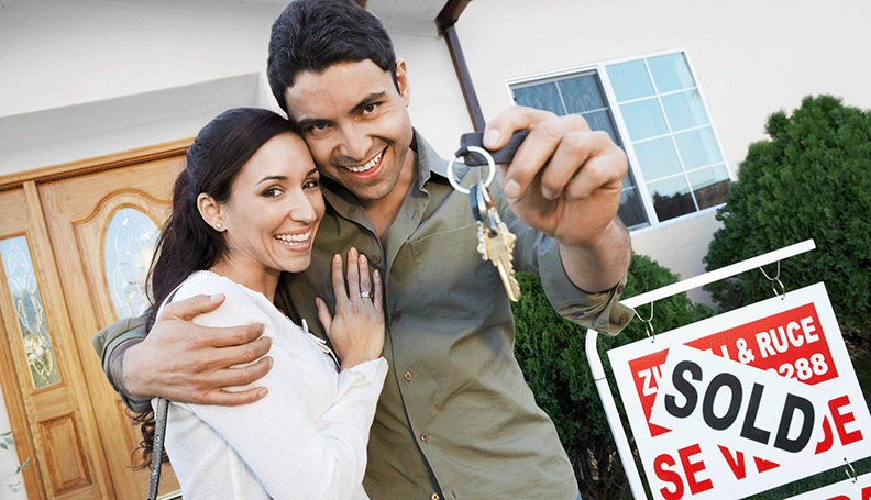 couple embracing and standing in front of sold sign as one holds up house keys to the viewer