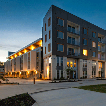 photograph of a newly-completed, five-story, mixed-use apartment building at dusk with its lights on