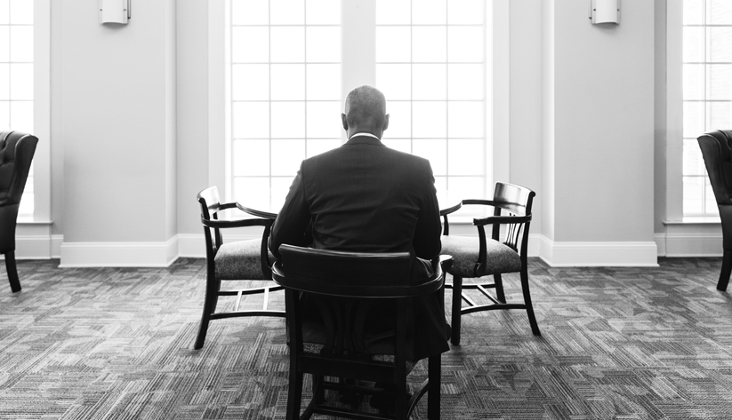 Portrait in black and white of Raphael Bostic sitting at a table alone with his back facing camera