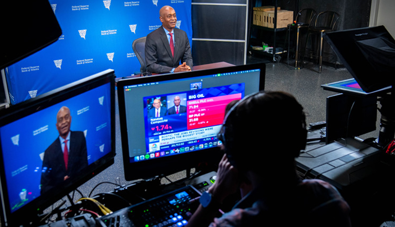 Raphael Bostic in background of sound studio being interviewed by CNBC anchor on the monitor in the foreground with recording technician