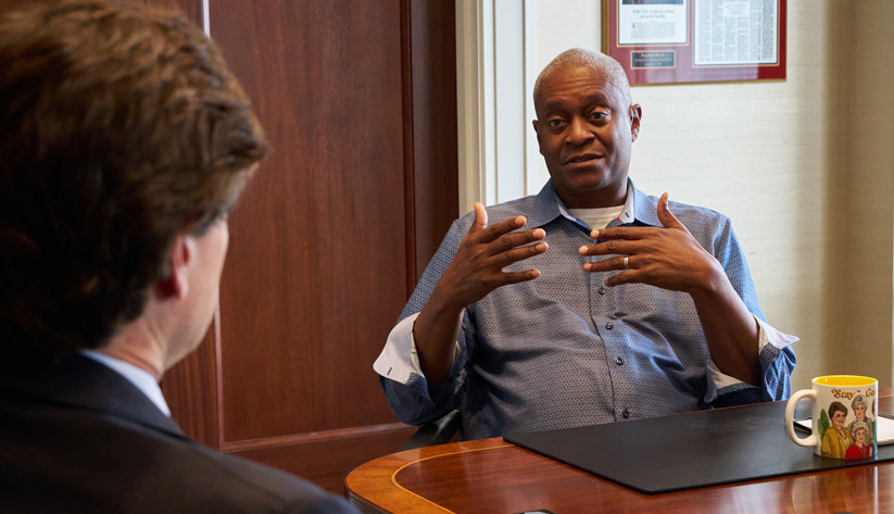 Raphael Bostic sitting at a table gesturing with hands speaking with man whose back is to the camera