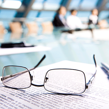 reading glasses over document in foreground in the background are out-of-focus people seated around an executive conference table