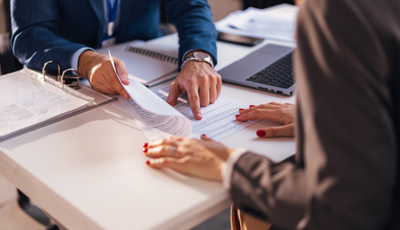 A person pointing to piece of paper on desk to other person sitting across from them
