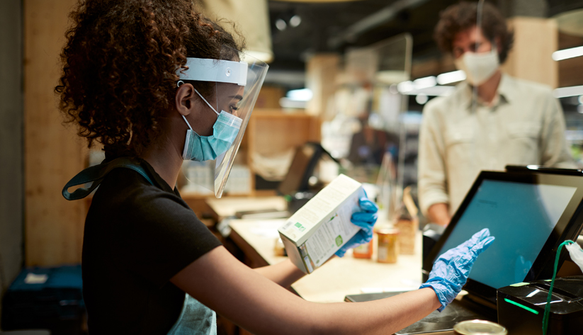cashier checking out item for customer behind protective screen wearing mask and gloves