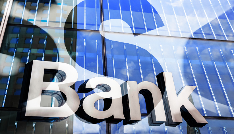 Side of a glass building with the word "Bank" in silver with blue dollar sign shadow on building