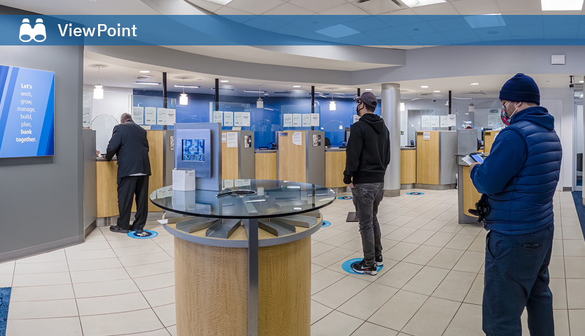 Bank customers standing on social distancing floor markers while waiting at teller stations in a modern branch with protective barriers.