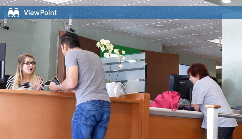 Bank teller assisting customer at counter while another customer conducts transaction at a separate station in a modern bank branch.