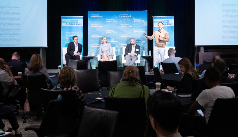 Long shot of group of people facing a stage with four panelists 