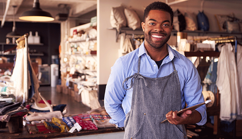 aproned man standing in foreground with upholstery materials and workspace in background