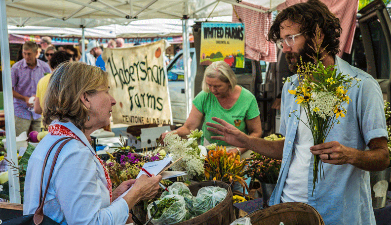 busy farmers market scene with two people speaking to each other in the foreground