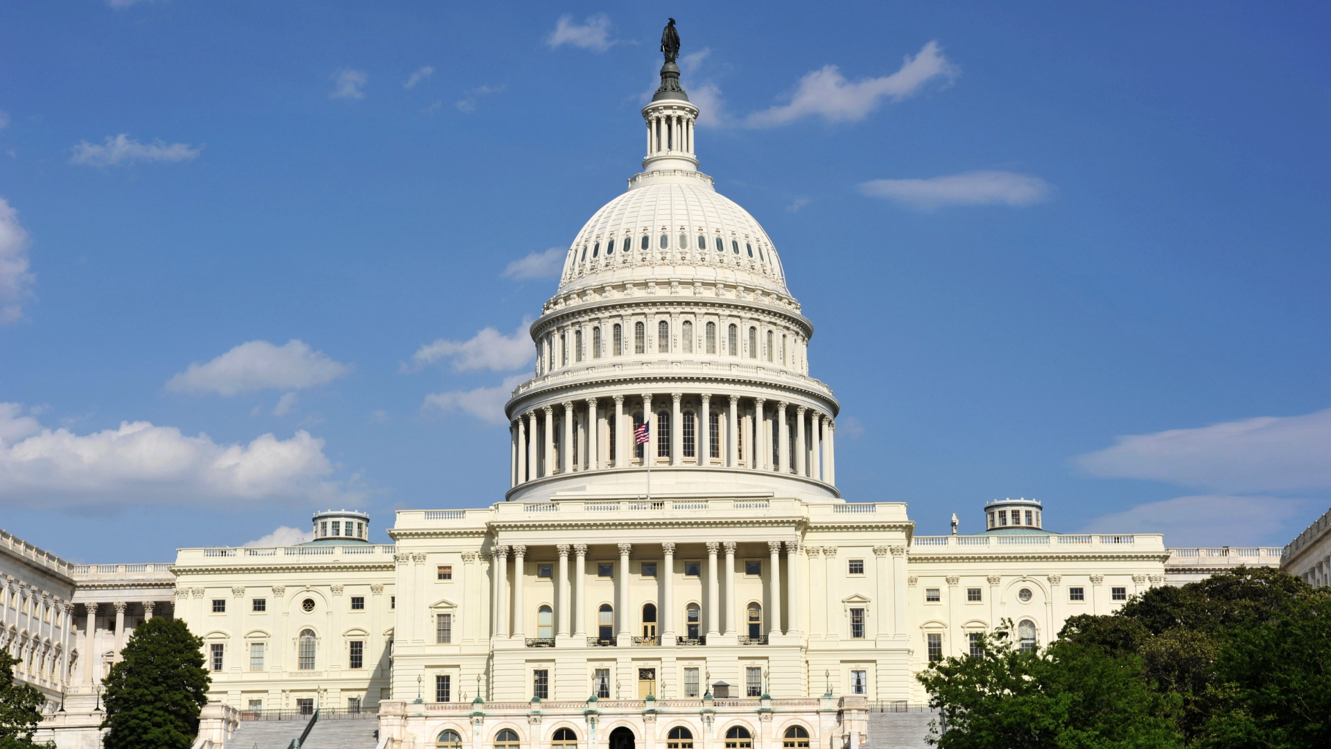 Photograph of the United States Capitol Building with its dome and the American flag in front of it with a backdrop of a partially cloudy daytime sky