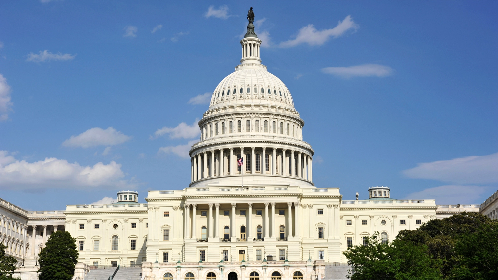 Photograph of the United States Capitol Building with its dome and the American flag in front of it with a backdrop of a partially cloudy daytime sky