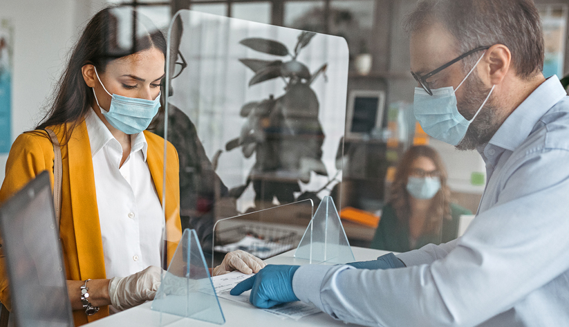 man and woman speaking with masks on behind plexy glass