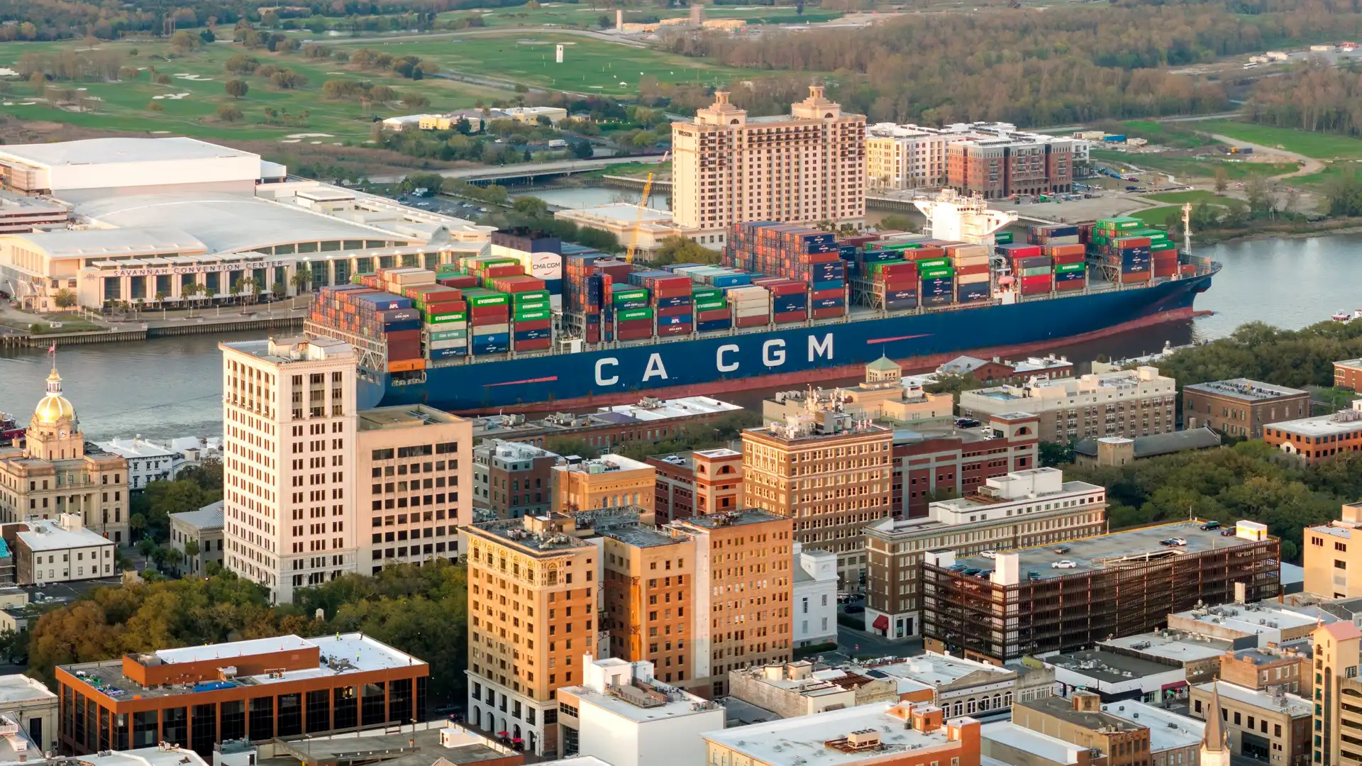 photograph of a heavily-loaded CMA CGM cargo container ship traveling behind Savannah skyline along Savannah River