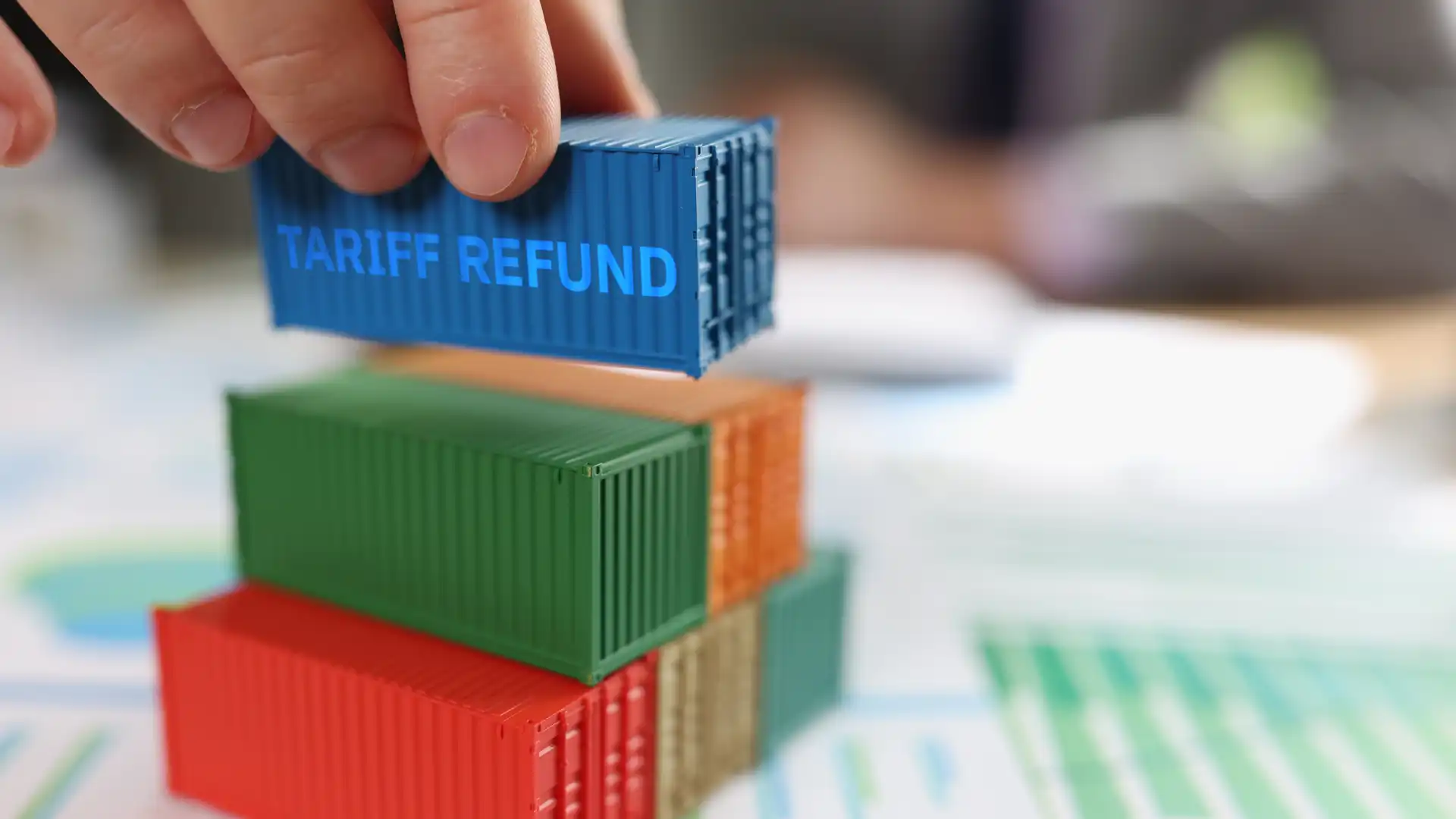 hand piling toy-sized shipping containers onto a pile of papers with charts and graphs with top container reading 'tariff refund' on its side