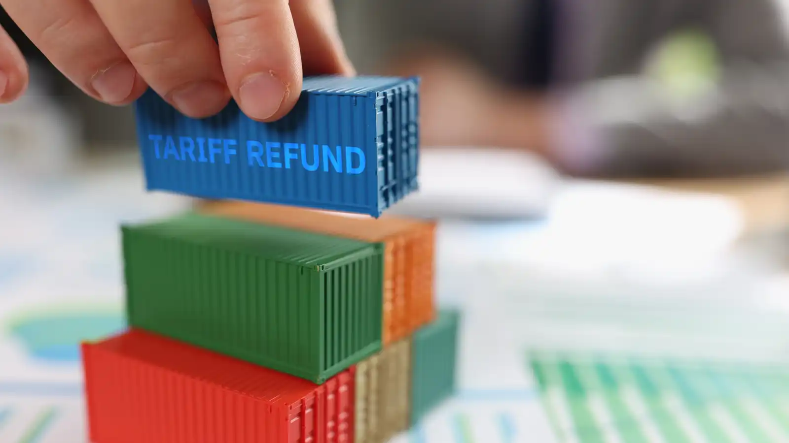 hand piling toy-sized shipping containers onto a pile of papers with charts and graphs with top container reading 'tariff refund' on its side