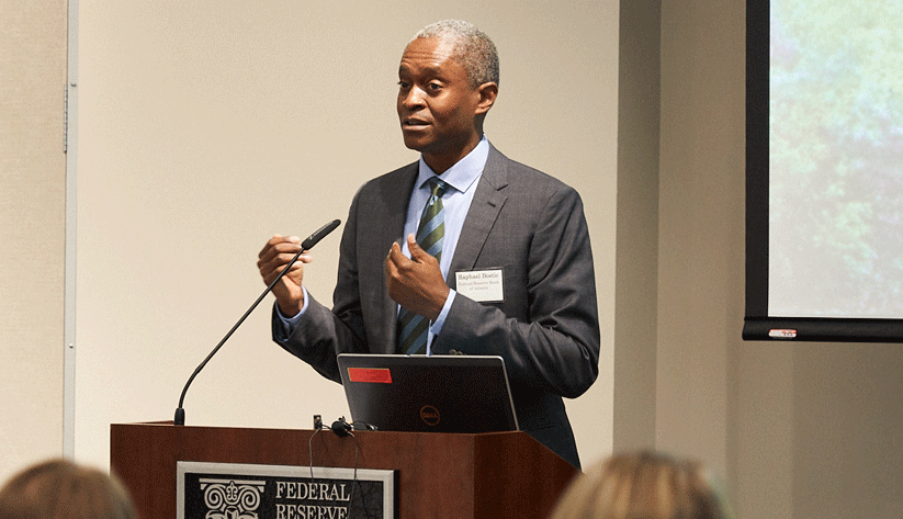 Raphael Bostic speaking at podium in front of screen and white wall with both hands up