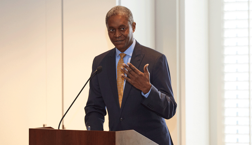Raphael Bostic speaking at a podium in front of a screen and white wall with left hand up.