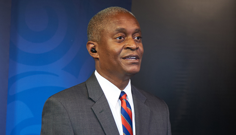 Photo of Raphael Bostic speaking in front of a blue curtain with the column logo