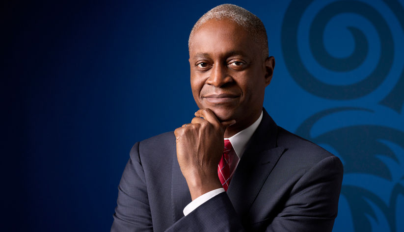 Portrait of Raphael Bostic in front of a blue screen with left hand at chin