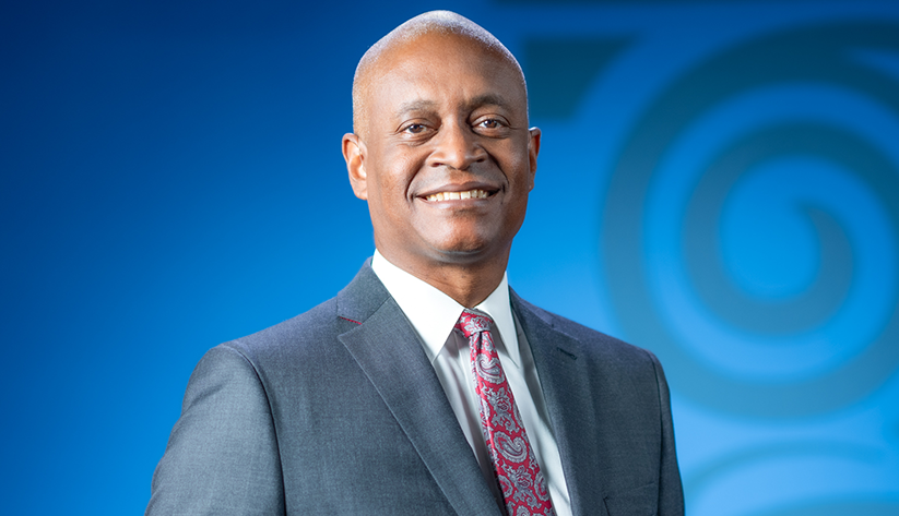 Portrait of Raphael Bostic in front of a blue screen with column logo