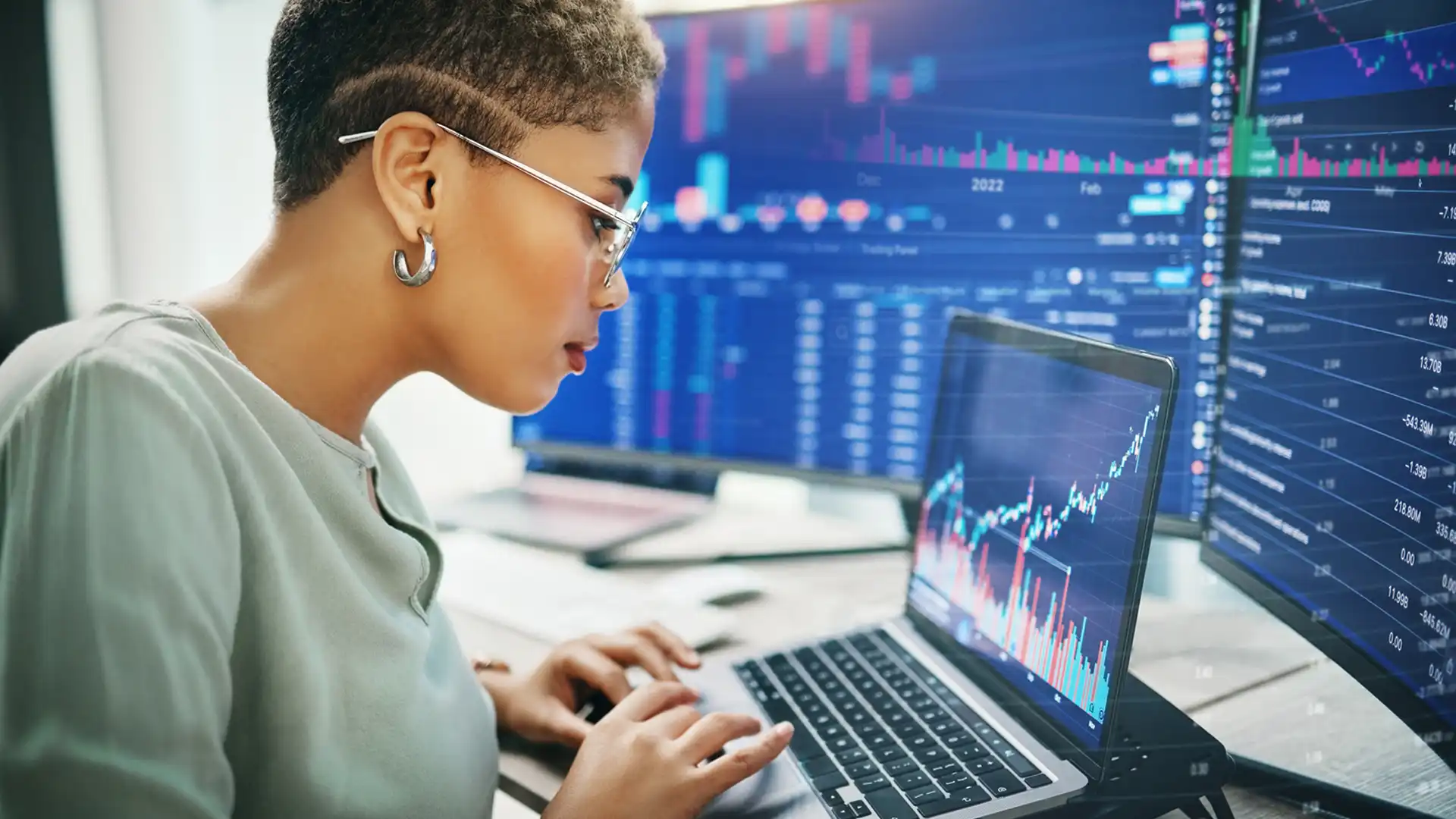 Woman working on data on laptop with two large monitors behind showing data