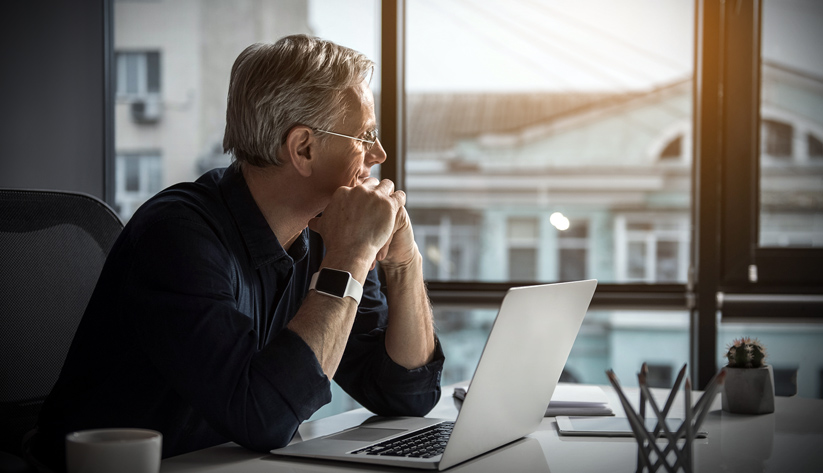 older man in front of computer at desk looking out window