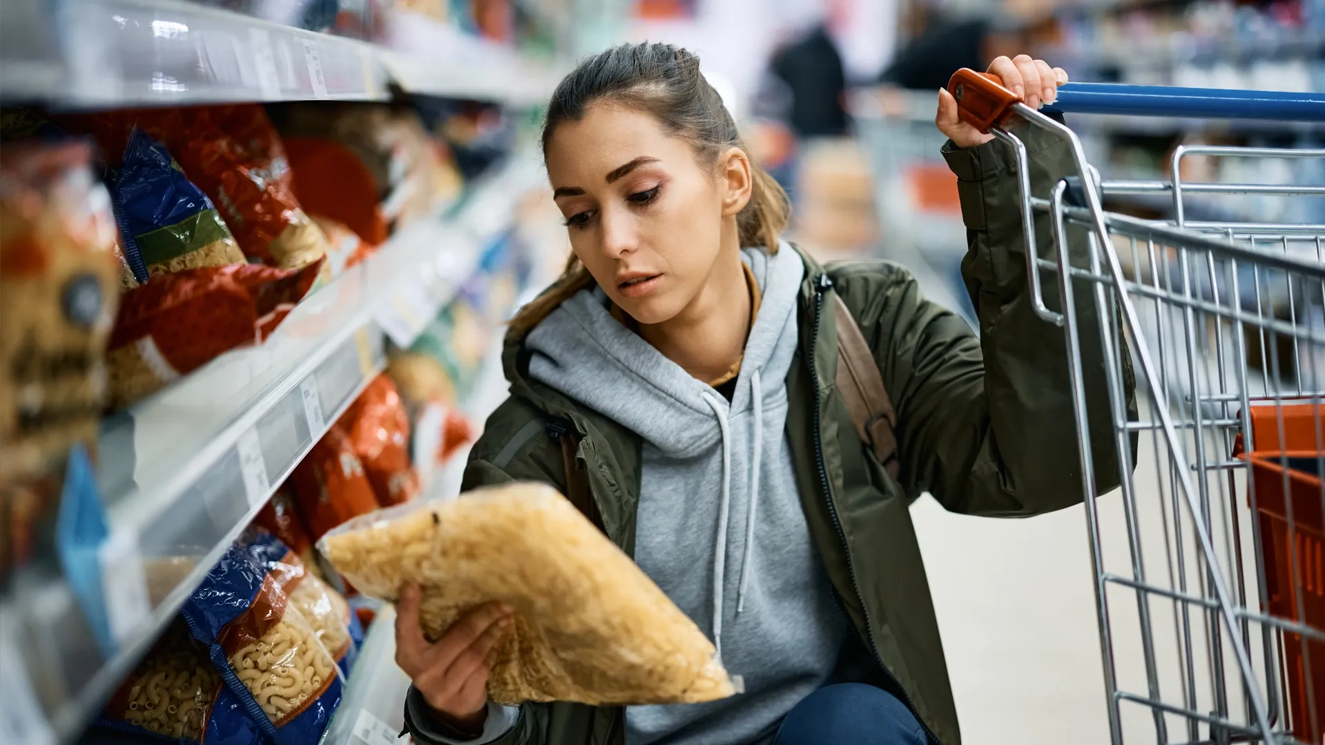 Woman in grocery store holding pasta in her right hand and left hand on a grocery cart