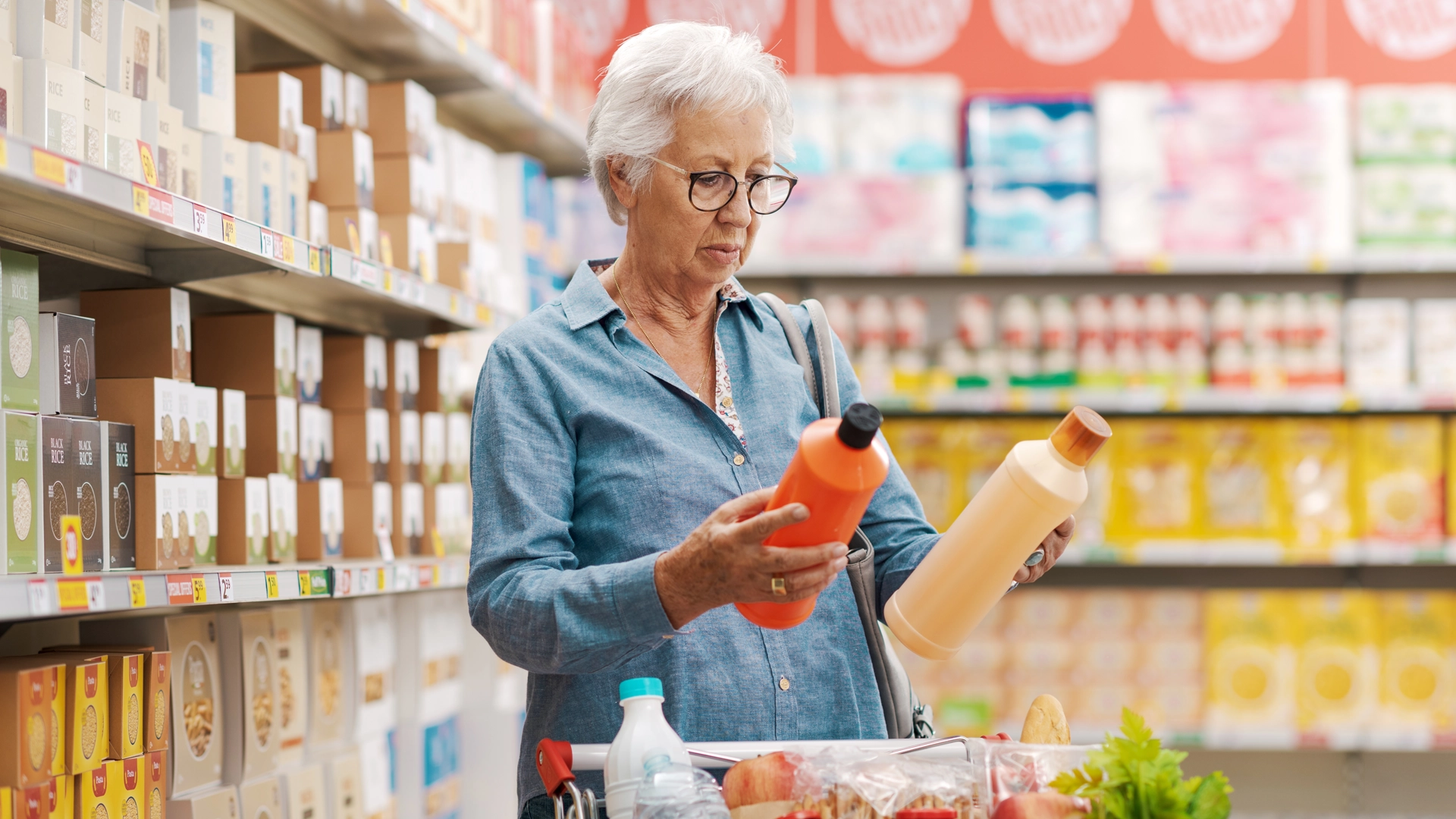 Woman wearing blue dress, glasses, with white hair comparing two grocery items